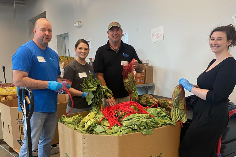 four people pose next to a large crate of fresh produce at a food bank